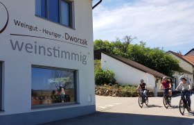 E-Bike Verleih, © weinstimmig Dworzak Drei Personen fahren mit Fahrrädern an einem Gebäude mit der Aufschrift 'Weingut - Heuriger - Dworzak weinstimmig' vorbei.