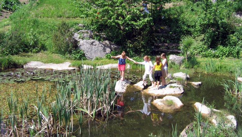 Idyllisches Natur-Biotop im Naturhotel Steinschalerhof, © Steinschalerhof Kinder spielen an einem Teich im Grünen mit Seerosen und Schilf.