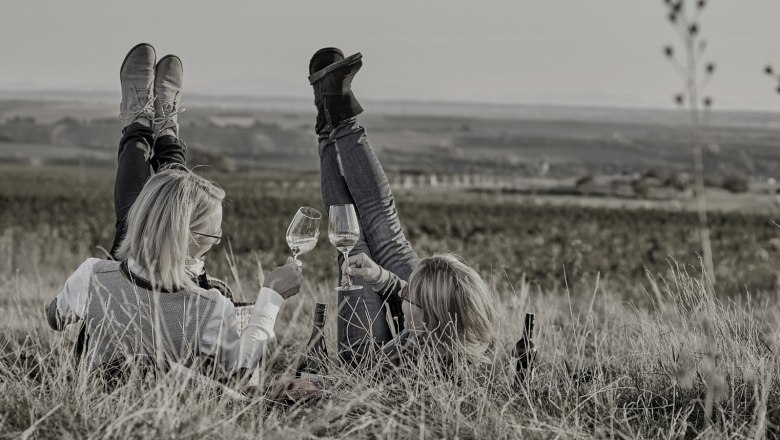 Genießen in Schrattenthal, © Weingut Hindler Zwei Personen liegen im Gras mit Weingläsern in der Hand, die Beine in die Luft gestreckt, vor einer weiten Landschaft.