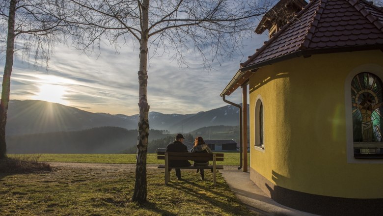 Kapelle - Ramswirt, © Gasthof Ramswirt Zwei Personen sitzen auf einer Bank neben einer kleinen Kapelle mit Blick auf die Berge bei Sonnenuntergang.