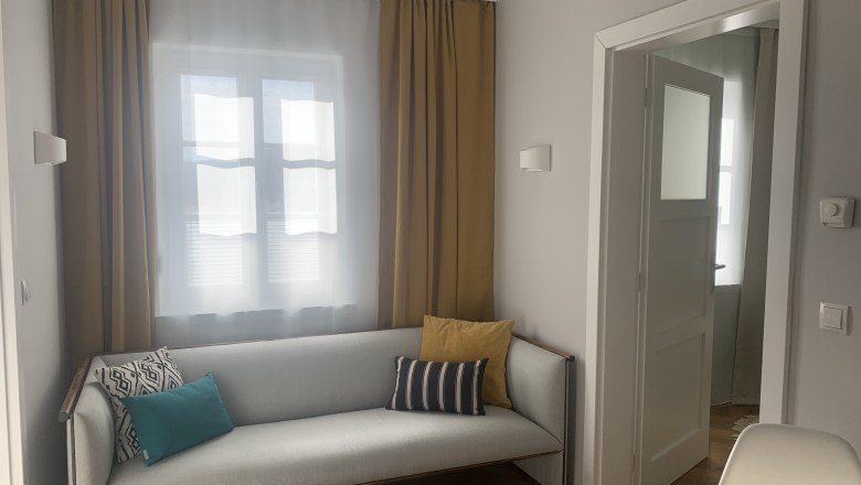 Apartment 1 "It's so cosy", © kremsoase Gemütliches Wohnzimmer mit Sofa und Kissen, gelben Vorhängen und offenem Türrahmen.