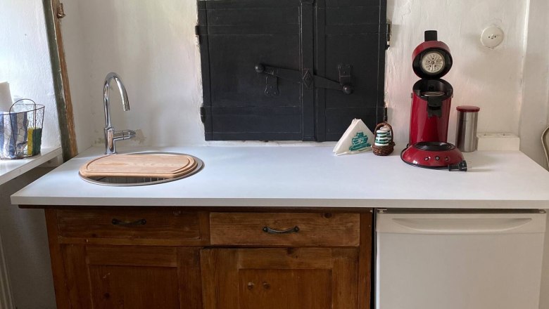 Küchenteil, © Familie Stagard Kleine Küche mit Holzschränken, weißer Arbeitsplatte, roter Kaffeemaschine und kleinem Kühlschrank.