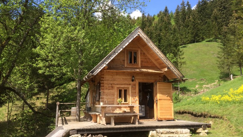 Holzknechthütte Hinterbreiteneben, © Johannes Hoyos Eine kleine Holzhütte in einer grünen, bewaldeten Landschaft mit blauem Himmel.