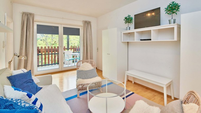 Top 2 - Wohnzimmer, © VP FeWo OG Modernes Wohnzimmer mit Sofa, Sessel, Glastisch und TV. Balkon mit Holzzaun im Hintergrund.