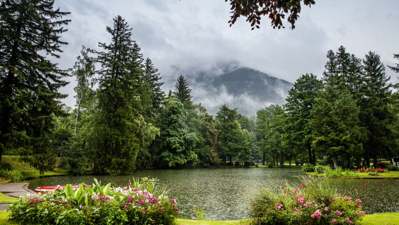 Kurpark Reichenau, © Wiener Alpen/Nadja Meister Ein malerischer Park mit einem Teich, umgeben von Bäumen und Blumen, vor einem nebligen Berg im Hintergrund.