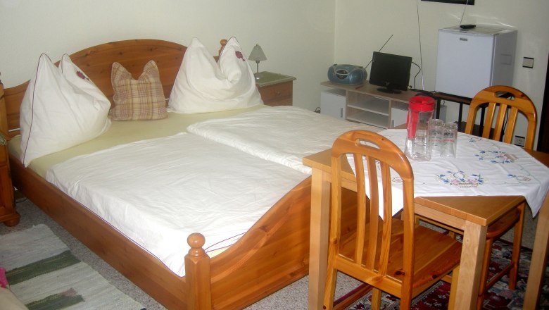Privatzimmer Gruber, © Siegfried Gruber Ein gemütliches Schlafzimmer mit Holzmöbeln, einem Doppelbett, einem kleinen Tisch mit Stühlen und einem Fernseher.