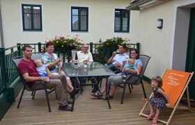 Familie Krottendorfer, © Familie Krottendorfer Eine Familie sitzt auf einer Terrasse an einem Tisch mit Getränken. Im Hintergrund sind Blumen und ein Gebäude zu sehen.