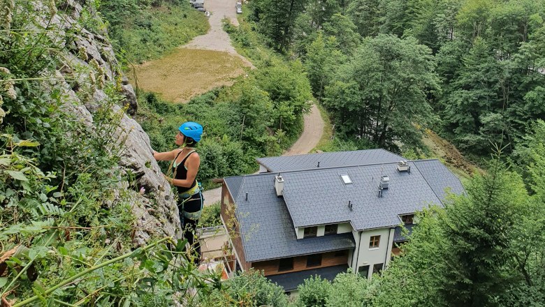Kletter-Yoga-Camp im Höllental, © Raufgeklettert - Petra Weisz Person klettert an einer Felswand mit Helm und Kletterausrüstung, Gebäude und Wald im Hintergrund.