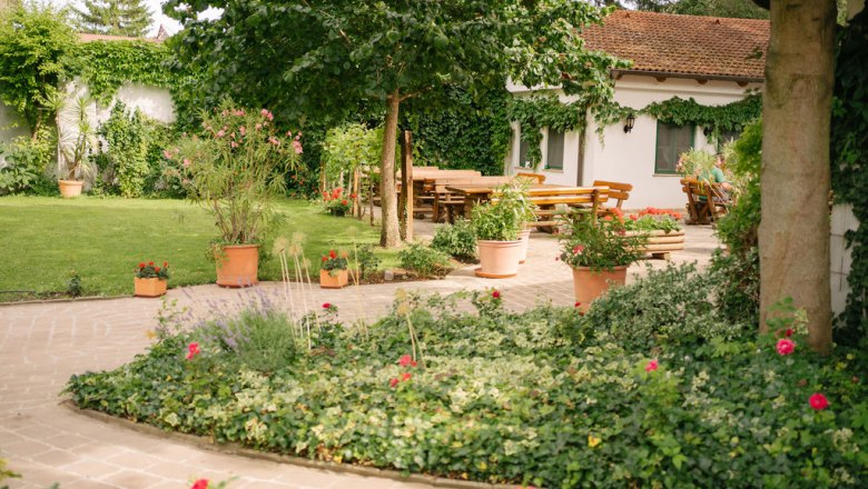 Garten, © Felix Sommer Ein gepflegter Garten mit Rasen, Blumenbeeten und Terrakotta-Töpfen. Im Hintergrund ein Haus mit Terrasse und Holztischen.