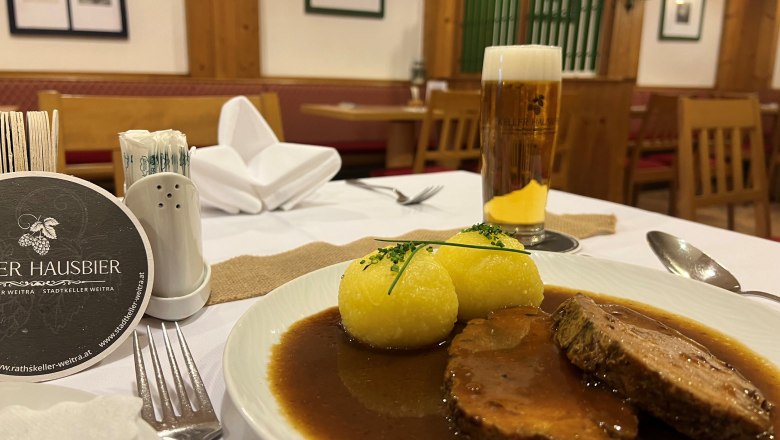 Schweinsbraten mit Erdäpfelknödel, © Matthias Bachofner Ein Teller mit Schweinsbraten und zwei Erdäpfelknödeln in einem Restaurant, daneben ein Glas Bier.