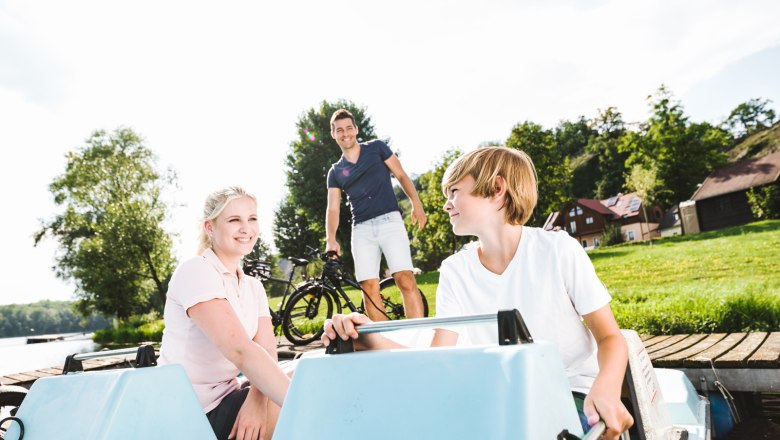 Entspannung im Freizeitzentrum Weitenegg, © NÖW_Stefan Fürtbauer Zwei Kinder in einem Tretboot, ein Mann mit Fahrrad im Hintergrund, grüne Wiese und Bäume.