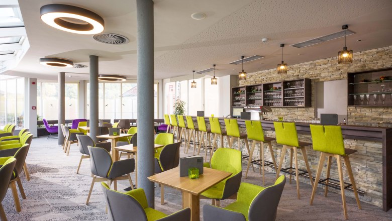 Café, © Hannes Dabernig Modernes Café mit grünen und grauen Stühlen, Holztischen und einer langen Bar mit Steinwand.