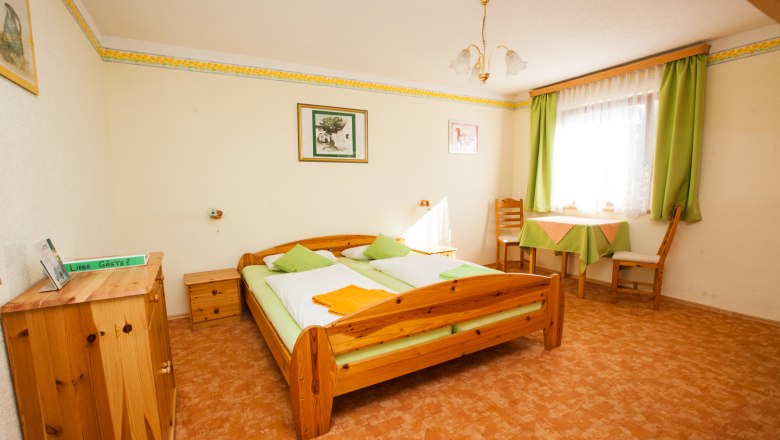 Gästezimmer, © Schneider Gemütliches Gästezimmer mit Holzmöbeln, Doppelbett, Tisch und Stühlen, grüne Vorhänge und Dekoration.