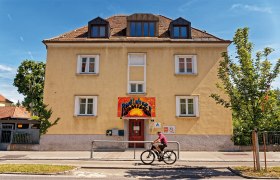 Aussen, © Christoph Sammer Gelbes Gebäude mit rotem Eingang und Graffiti-Schild. Ein Radfahrer fährt vorbei. Blauer Himmel und Bäume im Hintergrund.