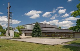 Pfarrkirche St. Paul, © Jürgen Übl Moderne Kirche mit Glockenturm und Kreuz, umgeben von Grünflächen und Bäumen, unter blauem Himmel mit Wolken.