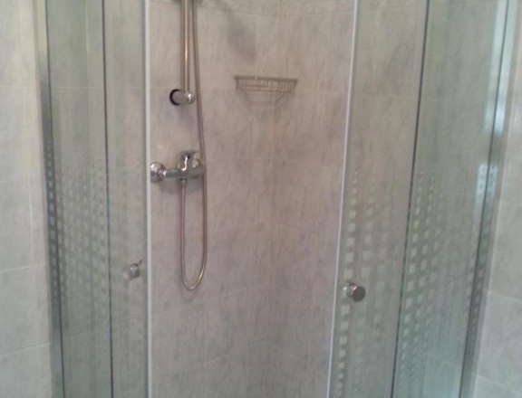 Badezimmer, © Haus Nova Eckdusche mit Glastüren und Duschkopf in einem gefliesten Badezimmer.