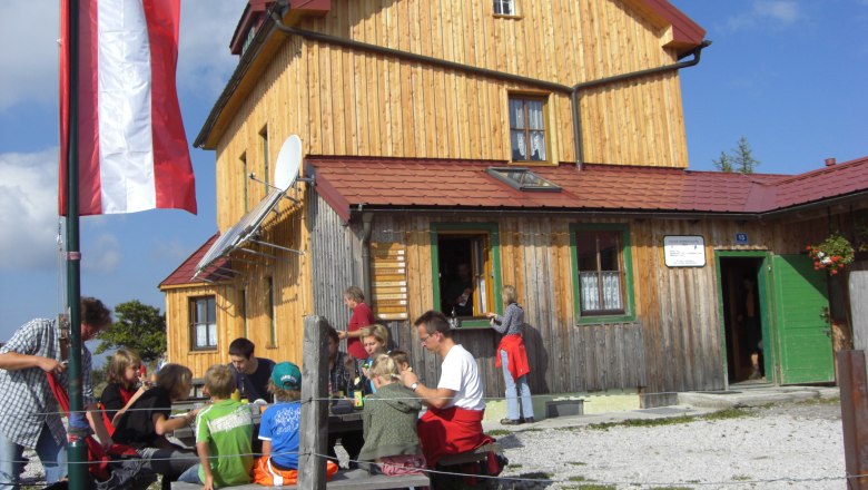 Julius Seitner Hütte, © Gemeinde Loich Eine Gruppe von Menschen sitzt vor einer Berghütte mit Holzfassade und österreichischer Flagge.