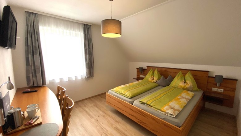 Doppelzimmer, © Trachsler Ein gemütliches Doppelzimmer mit einem Doppelbett, Schreibtisch, Stühlen und einem Fernseher an der Wand.