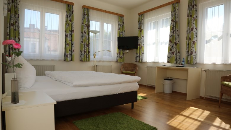 zimmeransicht, © Gerald Riedler Helles Zimmer mit Bett, Schreibtisch, Fernseher und mehreren Fenstern mit Vorhängen.