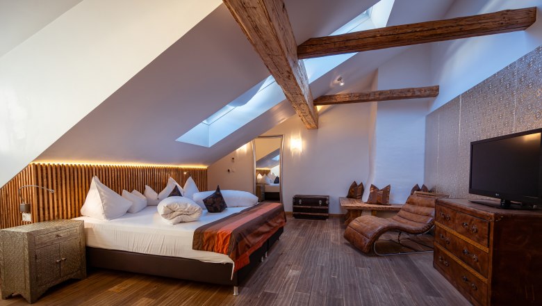 Hotel Landhaus Moserhof, © Niederösterreich Werbung / Maximilian Pawlikowsky Gemütliches Hotelzimmer mit Holzbalken, Dachfenstern, großem Bett und Ledersessel.