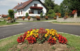 Gästehaus Reuberger, © Gästehaus Reuberger Ein weißes Haus mit roten Dachziegeln und Blumenschmuck, umgeben von bunten Blumenbeeten und grüner Landschaft.