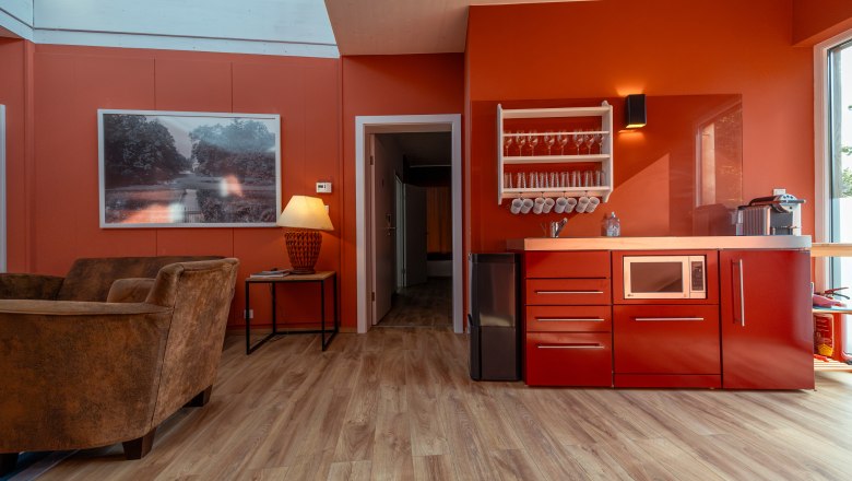 Grafenegg Cottages, © Niederösterreich Werbung / Maximilian Pawlikowsky Innenansicht eines modernen Wohnraums mit roten Wänden, einer roten Küchenzeile, braunen Sesseln und einem großen Fenster.