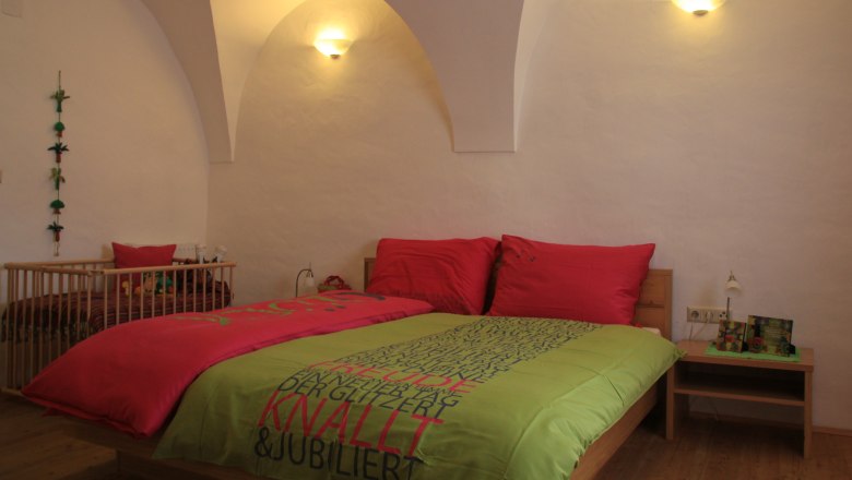 Gästezimmer, © Wolfgang Rieder Ein gemütliches Gästezimmer mit einem Doppelbett, bunten Bettbezügen und einem Babybett im Hintergrund. Die Wände sind weiß und gewölbt.