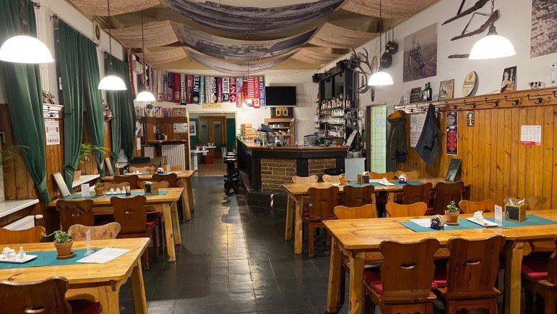 Gaststube, © Andreas Sinnhuber Gemütliche Gaststube mit Holztischen, Stühlen und Bar, dekoriert mit Fußballschals und rustikalen Elementen.