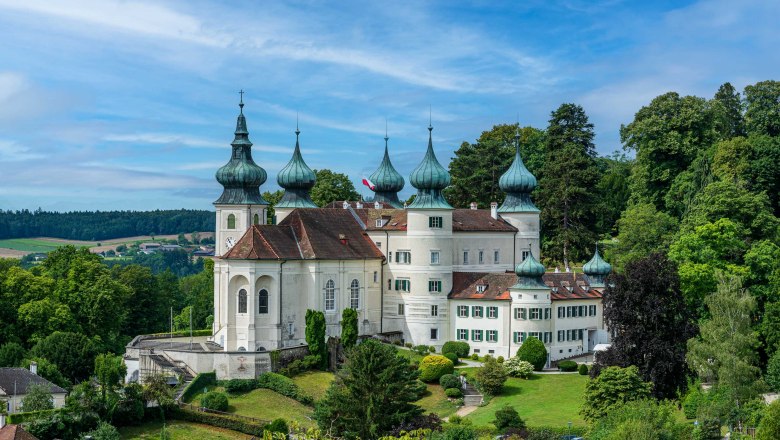 Schloss Artstetten mit Garten, © Schloss Artstetten/D. Mayrhofer Ein weißes Schloss mit mehreren Zwiebeltürmen, umgeben von grüner Landschaft und Bäumen, unter einem blauen Himmel.