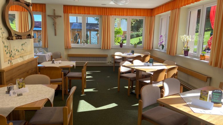 Speisesaal, © Wiener Alpen Ein heller Speisesaal mit Holztischen, Stühlen und großen Fenstern mit orangefarbenen Vorhängen.