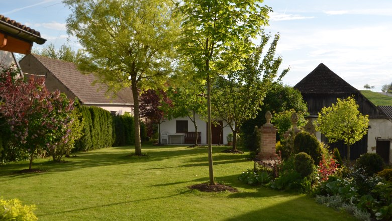 Garten, © Familie Herrmann Ein gepflegter Garten mit Rasen, Bäumen und Sträuchern, umgeben von Gebäuden und einem blauen Himmel im Hintergrund.
