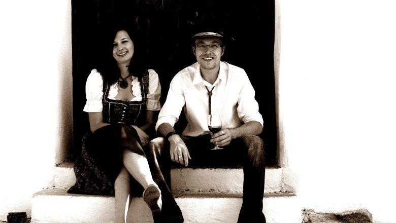 Winzer, © Weingut A+A Mayer Ein Mann und eine Frau sitzen auf einer Treppe, der Mann hält ein Weinglas.