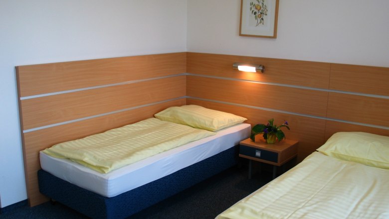 Zimmerbeispiel, © Gästehaus der LK-Technik Mold Ein einfaches Hotelzimmer mit zwei Einzelbetten, einem Nachttisch und einem Bild an der Wand.