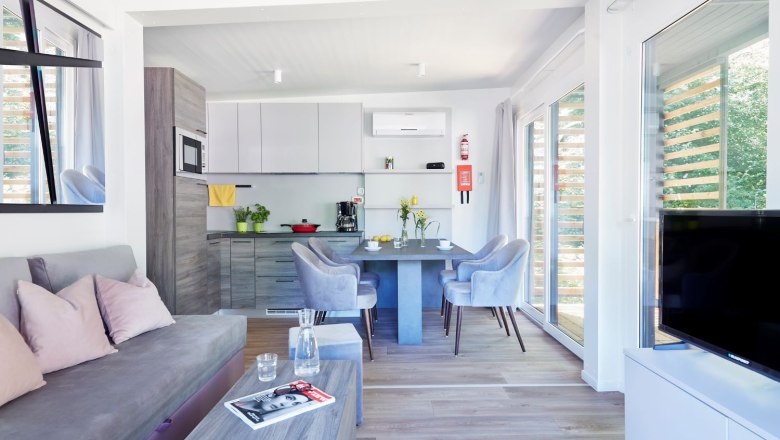 TDR-Mobilhome innen, © Campingplatz Tulln Mobilheim Wohnküche mit grauer Couch und weißer Küchenzeile sowie Flachbildschirm