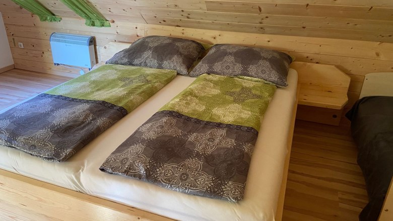 Ferienwohnung - Wohn und Schlafbereich in einem, © Wiener Alpen Doppelbett in einem Raum mit Holzverkleidung und grünen Vorhängen.