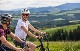 Aussichtsroute, © Wiener Alpen, Martin Fülöp Zwei Radfahrer mit Helmen fahren durch eine grüne Landschaft mit Hügeln im Hintergrund.