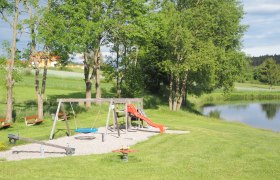 Spielplatz, © MG Sallingberg Spielplatz mit Schaukel, Rutsche und Wippe neben einem Teich in einer grünen Landschaft.