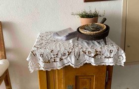 Detail im Wohnbereich, © Familie Stagard Ein kleiner Holztisch mit Spitzendecke, Pflanzen und einem gerahmten Bild an der Wand.