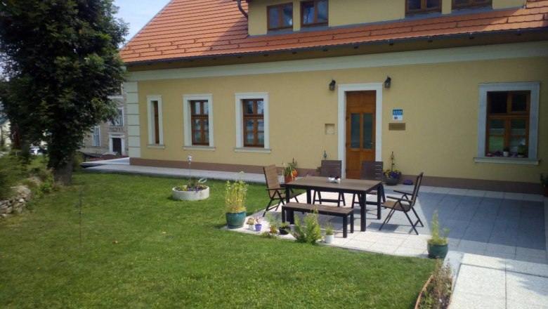 Terrasse, © Ildiko Davidne Nagy Ein gelbes Haus mit rotem Dach und Terrasse, umgeben von Rasen und Pflanzen.