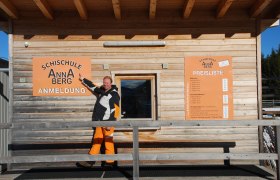 Schischule Annaberg, © NÖ-BBG Ein Mann zeigt auf ein Schild der Schischule Annaberg vor einem Holzgebäude.