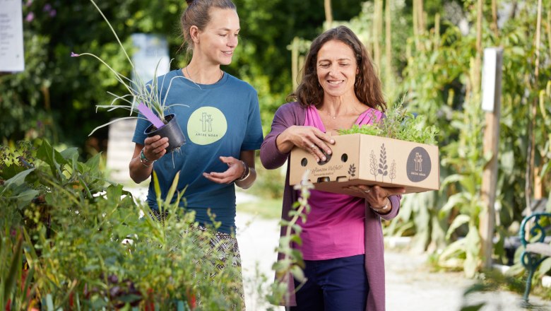 ARCHE NOAH, © ARCHE NOAH_Rupert Pessl Zwei Frauen in einem Garten, eine trägt eine Kiste mit Pflanzen, die andere hält einen Blumentopf. Beide lächeln und unterhalten sich.
