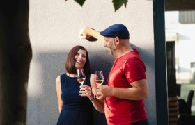 Sigrid und Christoph Schleinzer, © Astrid Bartl Ein Mann und eine Frau stehen lächelnd an einer Wand, halten Weingläser und sind von Blättern beschattet.
