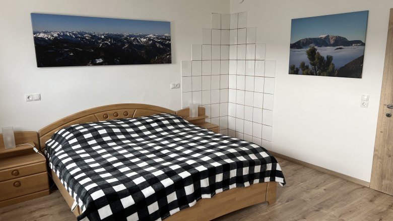 Doppelbett, © Anton Sommer Schlafzimmer mit Doppelbett, kariertem Bettüberwurf, Holzmöbeln und Berglandschaftsbildern an der Wand.