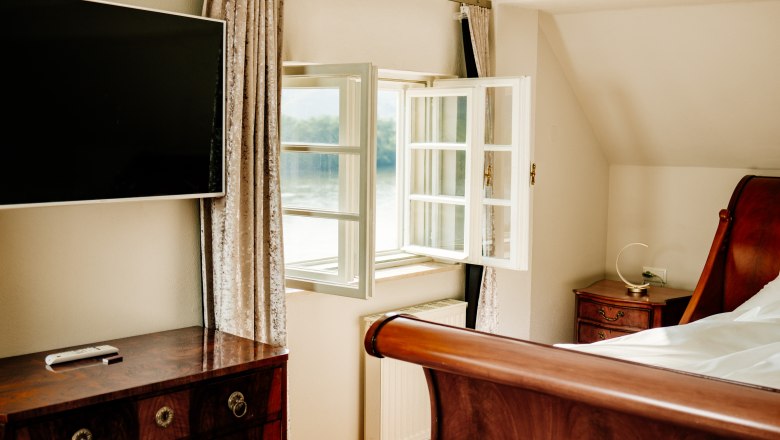 villa_schonthal-25_2, © Julius Hirtzberger Ein elegantes Schlafzimmer mit einem offenen Fenster, einem Flachbildfernseher, einem Holzbett und antiken Möbeln.