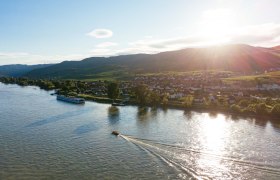 ahoi-wachau-Luftaufnahme, © NÖW_Daniel Gollner Luftaufnahme der Wachau mit Fluss, Booten und Dorf im Sonnenuntergang.