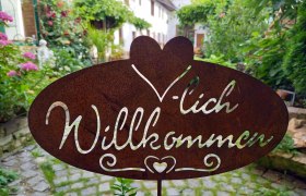 Herzlich Willkommen, © Familie Fleischmann Metallschild mit der Aufschrift 'Herzlich Willkommen' in einem Garten.