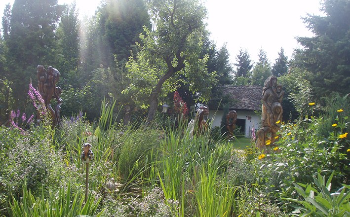 SkulpturNaturgarten, © Harry Raab Ein üppiger Garten mit Skulpturen, Bäumen und einem kleinen Gebäude im Hintergrund.