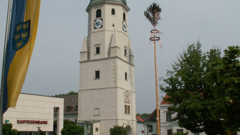 Fischamend, © Stadtgemeinde Fischamend Ein Kirchturm mit Uhr und grünem Dach in Fischamend, daneben ein Maibaum und eine Raiffeisenbank-Filiale.