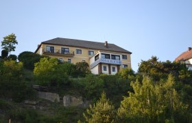 Außenansicht1, © Gasthof Donaublick Ein gelbes Gebäude mit der Aufschrift 'Donaublick' auf einem Hügel, umgeben von Bäumen und blauem Himmel.