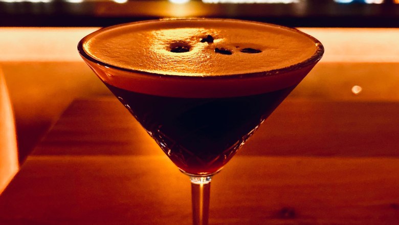 Bargetränk, © Markus Jokesz Ein Cocktailglas mit dunklem Getränk auf einem Holztisch, im Hintergrund eine unscharfe Bar.